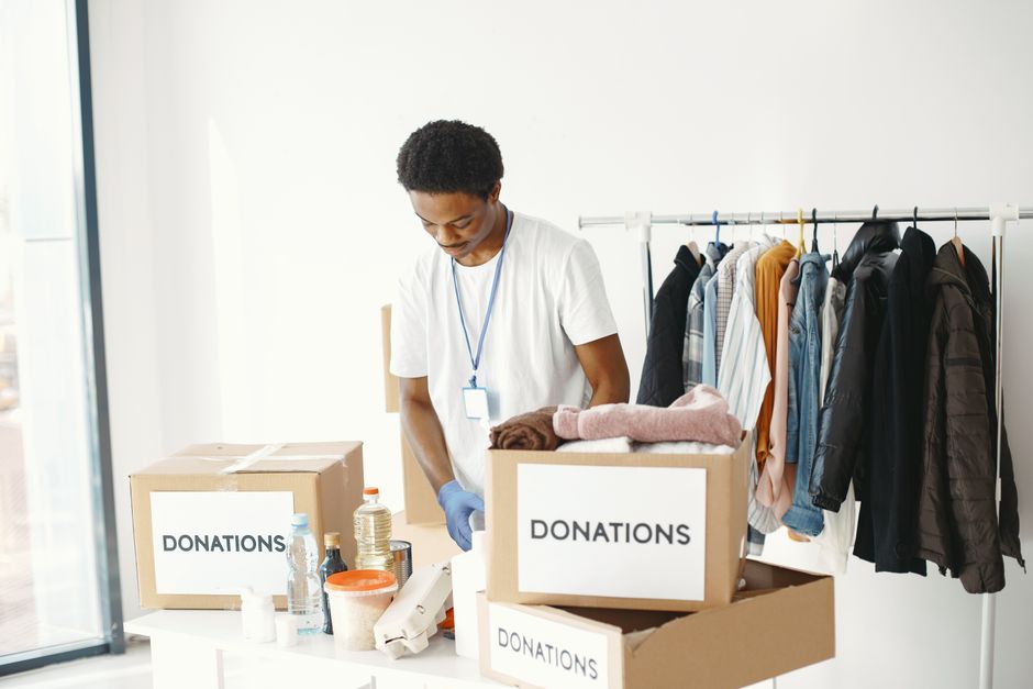 A person holding a cardboard sign with the word 'Donation' written in black ink, standing behind a table with several transparent plastic donation boxes. The donation boxes contain folded clothes and 