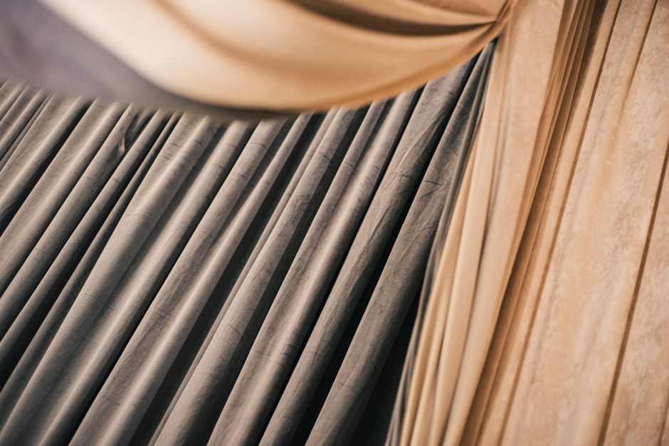 Close-up of light-colored, semi-transparent curtains made of fabric with a subtle checkered pattern, hanging neatly in a domestic setting. The curtains are well-maintained and appear clean, allowing d